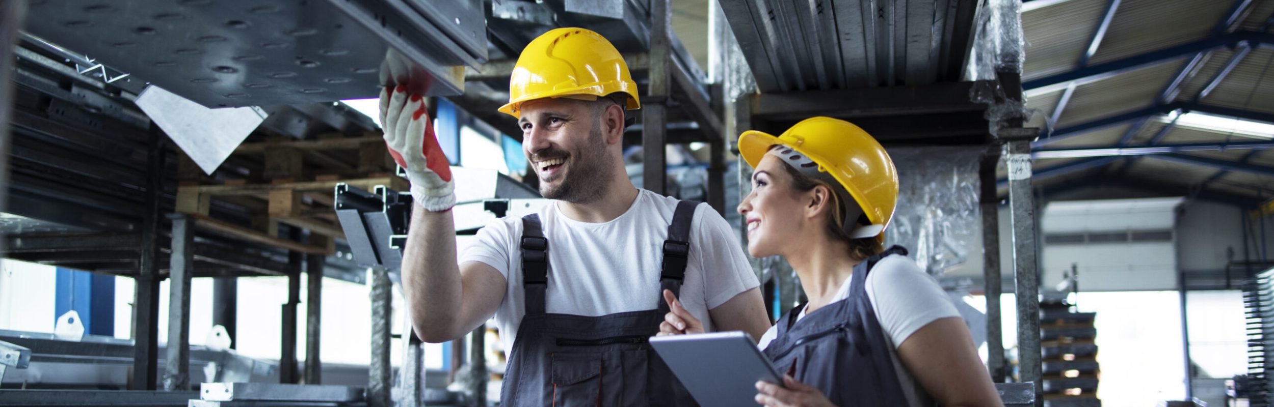 Factory workers checking inventory with tablet computer in industrial warehouse full of metal parts.